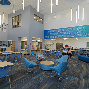 A modern elementary school media center with an open-concept design. The space features high ceilings, large windows, and contemporary lighting fixtures. Blue chairs and round tables provide flexible seating, while bookshelves line the walls. A motivational quote on the wall reads, "Believe in yourself and follow your dreams—you can achieve great things." A reading nook with soft seating is present, creating a welcoming environment for students.